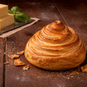 A golden, spiraled pastry on a dark wooden table with a block of butter and basil in the background.