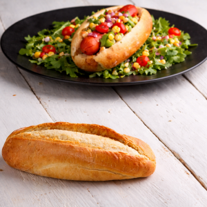Fresh baguette on a light wooden surface with a colorful hot dog plate in the background on a dark plate and greens nearby.