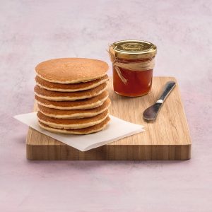 Stack of golden pancakes on a wooden board with a jar of maple syrup and a knife nearby.