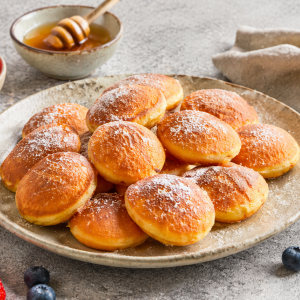 Plate of golden brown, powdered-sugar dusted mini buns on a rustic platter with honey and berries in the background.