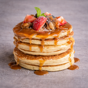Stack of fluffy pancakes with caramel sauce drizzling down the sides, topped with strawberries and berries with a mint leaf.