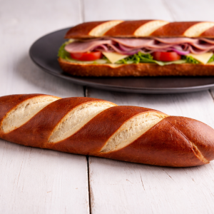Crusty baguette with diagonal slashes on a white wooden table; a ham and cheese sandwich on a plate in the background.