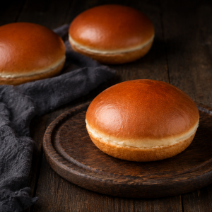 Three glossy brioche buns sit on a round wooden board on a dark table with a cloth nearby.